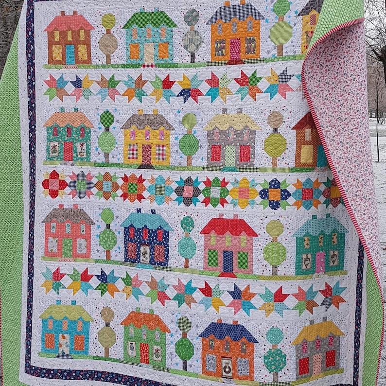 May include: A handmade quilt featuring a repeating pattern of colorful houses and trees on a white background. The quilt has a patchwork design with star and floral accents. The edges are finished with a green border and a floral patterned backing.