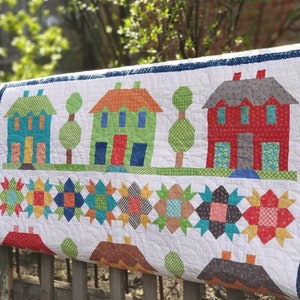 May include: A handmade quilt featuring a village scene with colorful houses, trees, and star patterns. The houses have orange, green, and red roofs, with blue, green, and red doors. The quilt is made of various patterned fabrics.