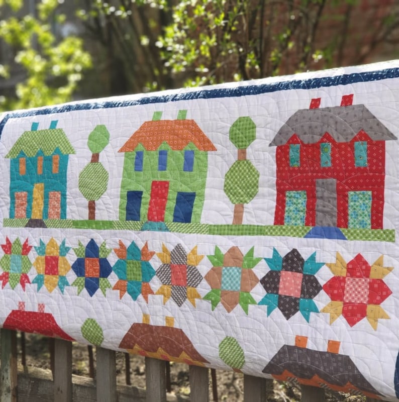 May include: A handmade quilt featuring a village scene with colorful houses, trees, and star patterns. The houses have orange, green, and red roofs, with blue, green, and red doors. The quilt is made of various patterned fabrics.