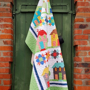May include: A colorful handmade quilt featuring a patchwork design of houses, stars, and geometric shapes. The quilt has a white background with green, blue, red, and yellow accents. The quilt is hanging on a green door, with a brick wall in the background.