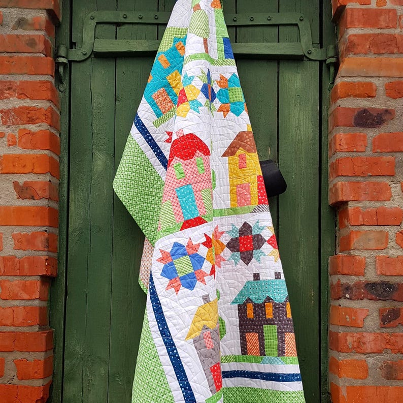May include: A colorful handmade quilt featuring a patchwork design of houses, stars, and geometric shapes. The quilt has a white background with green, blue, red, and yellow accents. The quilt is hanging on a green door, with a brick wall in the background.