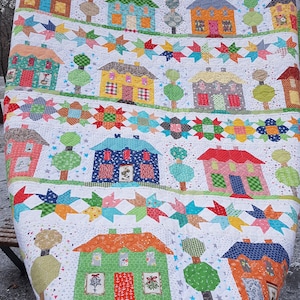 May include: A colorful patchwork quilt featuring rows of houses, trees, and star patterns. The houses are various colors, including brown, blue, and orange, with detailed windows and doors. The quilt has a white background with a green border.