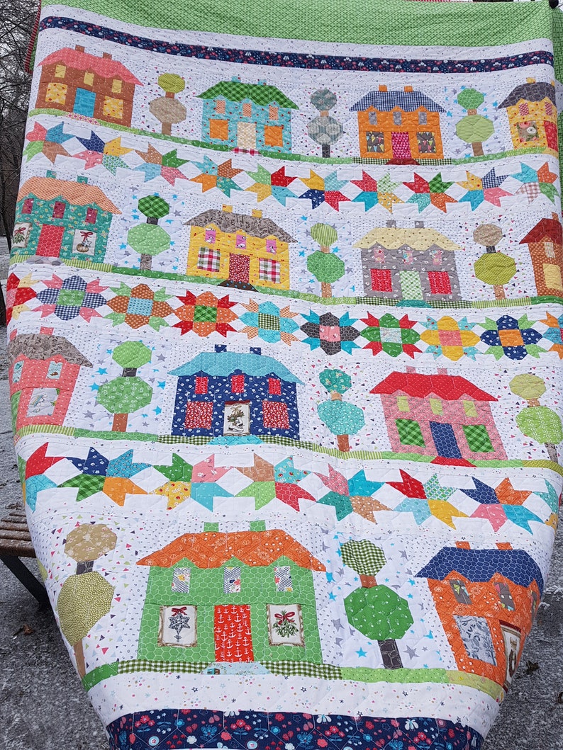 May include: A colorful patchwork quilt featuring rows of houses, trees, and star patterns. The houses are various colors, including brown, blue, and orange, with detailed windows and doors. The quilt has a white background with a green border.