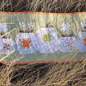 May include: A green and orange quilted table runner with a row of five white pumpkins with different coloured star and pinwheel designs.