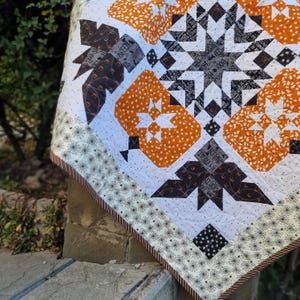 May include: A Halloween-themed quilt with a white background and orange, black, and gray patchwork squares. The quilt features a variety of Halloween-themed patterns, including pumpkins, bats, and spiders. The quilt is edged with a black and white spiderweb pattern.
