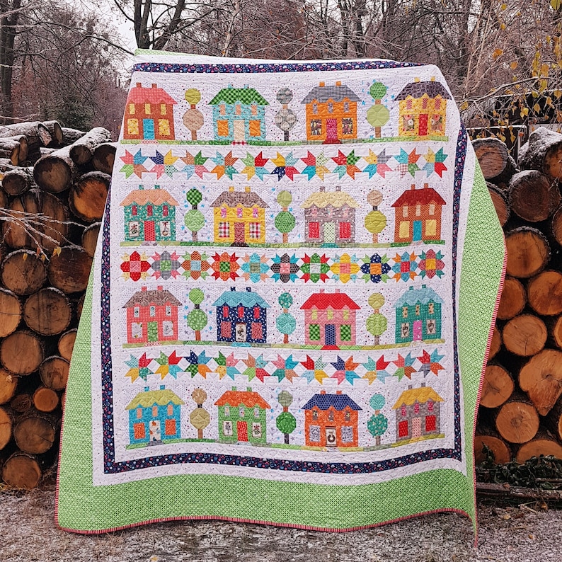 May include: A colorful quilt featuring rows of houses, trees, and star patterns. The houses have various colors and designs, with a white background. The quilt has a green border and is displayed outdoors.