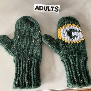May include: A pair of dark green knit mittens with a speckled texture and ribbed cuffs. One mitten features a white letter G outlined in yellow on the back. A small white sign above the mittens displays the black text ADULTS in capital letters.