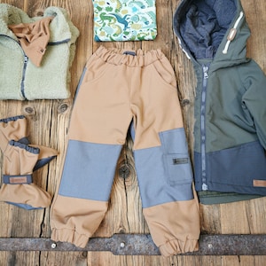 Może przedstawiać: Zestaw odzieży dziecięcej, w skład którego wchodzi zielona kurtka polarowa, brązowe i niebieskie spodnie, zielono-biała koszula z długim rękawem z nadrukiem dinozaura, zielona kurtka z kapturem z czarną podszewką oraz para szarych rękawic.