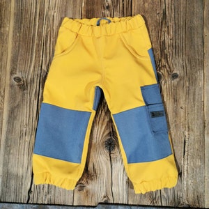 Może przedstawiać: Żółto-niebieskie spodnie dla malucha z elastyczną talią i kieszeniami. Spodnie mają niebieskie panele na kolanach i małą kieszeń na prawej nogawce. Mankiety są elastyczne.