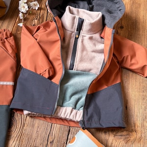 Op de afbeelding: Een kinderjasje in tinten oranje, grijs en lichtblauw, met een grijze capuchon en een lichtbruine binnenlaag. De jas heeft een ritssluiting en wordt gepresenteerd op een houten oppervlak. De jas maakt deel uit van een set.