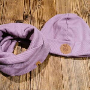 Könnte beinhalten: Eine lila gerippte Strickmütze und ein passender Infinity-Schal. Die Mütze hat einen braunen Leder-Patch mit einem Vogelmotiv.
