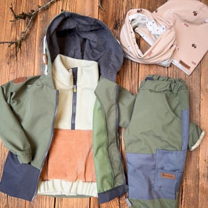 Könnte beinhalten: Ein Outdoor-Bekleidungsset für Kinder in Grün-, Grau- und Orangetönen, bestehend aus einer Kapuzenjacke, einer Hose, einem Halstuch und einer Mütze. Die Jacke hat ein Colorblock-Design mit einem Cord-Einsatz. Die Mütze hat einen Traktor-Aufdruck.