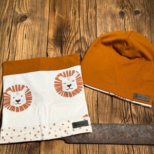 May include: An orange beanie and a white and orange lion-themed neck warmer set. The neck warmer features two lion faces and a dotted pattern. The beanie has a white trim and a small label.