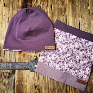 Könnte beinhalten: Eine lilafarbene Mütze mit einem floralen Futter und einem braunen Lederlabel mit der Aufschrift "Handmade". Die Mütze wird mit einem lilafarbenen und weißen floralen Halstuch mit einem gestreiften Rand kombiniert.