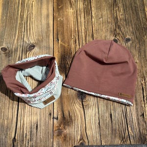 Puede incluir: Un conjunto a juego de un gorro color malva y un calentador de cuello. El gorro y el calentador de cuello están hechos de una tela malva lisa con un ribete estampado floral. El calentador de cuello tiene un interior a rayas. Ambos artículos tienen una pequeña etiqueta marrón.
