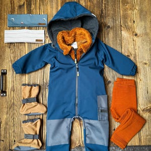 Peut inclure: Un ensemble pour bébé comprenant une combinaison à capuche bleue avec doublure grise et intérieur en polaire marron, un legging orange, deux bandeaux et des chaussons beiges. La combinaison a une fermeture éclair et des détails gris, sur une surface en bois.