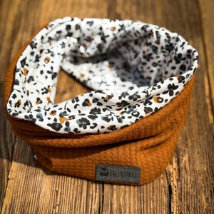 Könnte beinhalten: Ein rostfarbener Schlauchschal aus Rippstrick mit floral gemustertem Futter. Das Futter hat einen weißen Hintergrund mit schwarzen, braunen und blauen floralen Akzenten. Ein kleines, dunkelgraues Etikett ist auf den Schal genäht.