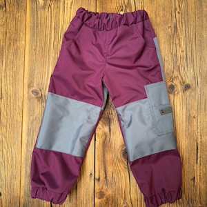 Może przedstawiać: Śliwkowe spodnie dziecięce z szarymi panelami na nogawkach i szarą kieszenią. Spodnie mają elastyczną talię i marszczone kostki. Tło to drewniana powierzchnia.