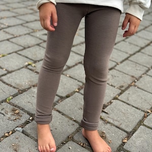 Könnte beinhalten: Braune Leggings mit einer feinen, horizontal gerippten Textur. Die Leggings sind lang geschnitten, haben eine körpernahe Passform und werden mit einem cremefarbenen Langarm-Sweatshirt kombiniert, das bis zum Oberschenkelbereich reicht.