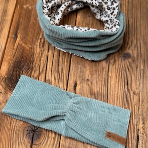 May include: A teal blue corduroy headband with a brown leather tag that says "DRECKSPATZ". The headband is folded in half and has a twisted knot in the center.