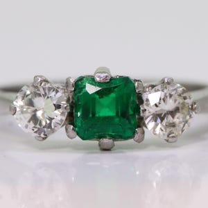May include: A three-stone ring featuring a square-cut emerald center stone flanked by two round-cut diamonds. The ring is set in a silver-colored band, showcasing the vibrant green and clear gemstones.