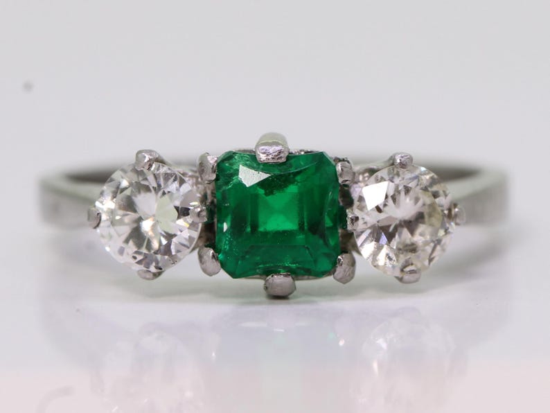 May include: A three-stone ring featuring a square-cut emerald center stone flanked by two round-cut diamonds. The ring is set in a silver-colored band, showcasing the vibrant green and clear gemstones.