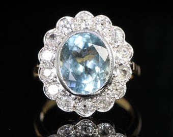 May include: An oval-shaped, light blue gemstone ring, surrounded by a halo of smaller, clear gemstones. The ring's setting is gold-colored, and the gemstones are set in a silver-colored metal. The ring is displayed against a black background.