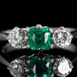 May include: A platinum ring featuring a square-cut emerald center stone flanked by two round-cut diamonds. The ring is set against a black background, highlighting the sparkle of the gemstones. A classic three-stone design.