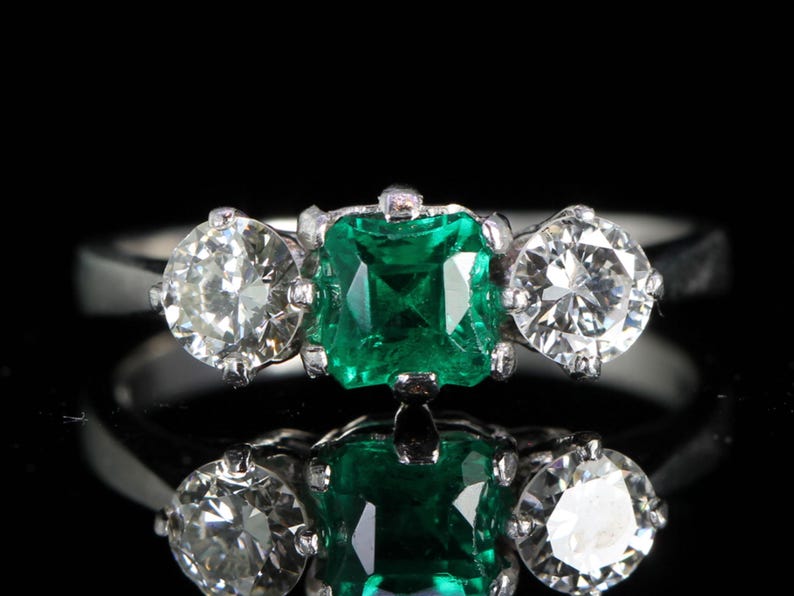 May include: A platinum ring featuring a square-cut emerald center stone flanked by two round-cut diamonds. The ring is set against a black background, highlighting the sparkle of the gemstones. A classic three-stone design.