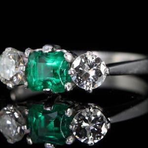 May include: A platinum ring featuring a central emerald-cut emerald flanked by two round-cut diamonds. The emerald is a vibrant green, and the diamonds sparkle. The ring is set against a black background.