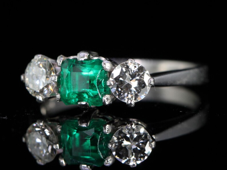 May include: A platinum ring featuring a central emerald-cut emerald flanked by two round-cut diamonds. The emerald is a vibrant green, and the diamonds sparkle. The ring is set against a black background.