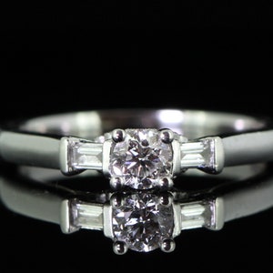 Może przedstawiać: Srebrny pierścionek zaręczynowy z centralnym diamentem okrągłym i dwoma diamentowymi bagietkami po obu stronach.