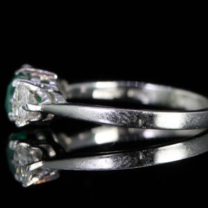 May include: A silver ring with a central oval-cut green gemstone, flanked by two smaller, triangular-cut clear gemstones. The ring is set against a black background, highlighting its reflective surface and the sparkle of the stones.