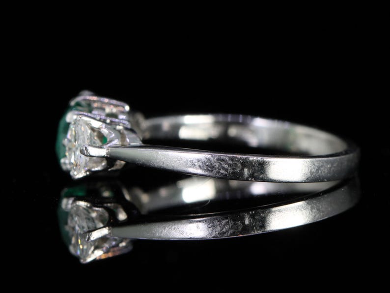 May include: A silver ring with a central oval-cut green gemstone, flanked by two smaller, triangular-cut clear gemstones. The ring is set against a black background, highlighting its reflective surface and the sparkle of the stones.