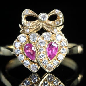 Op de afbeelding: Een gouden ring met een strik en twee hartvormige elementen. Elk hart bevat een roze edelsteen omringd door witte edelstenen. De strik en harten zijn versierd met witte edelstenen, wat een sprankelend effect creëert.