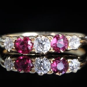 May include: A gold ring with a sequence of gemstones. The ring showcases a central diamond, with vivid red rubies and smaller diamonds on either side. The gemstones are set in a gold band, creating a classic and elegant design.