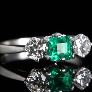 May include: A three-stone ring with a central emerald-cut green gemstone flanked by two round, brilliant-cut diamonds. The ring is set in a silver band, reflecting light against a black background. The ring's design is classic and elegant.