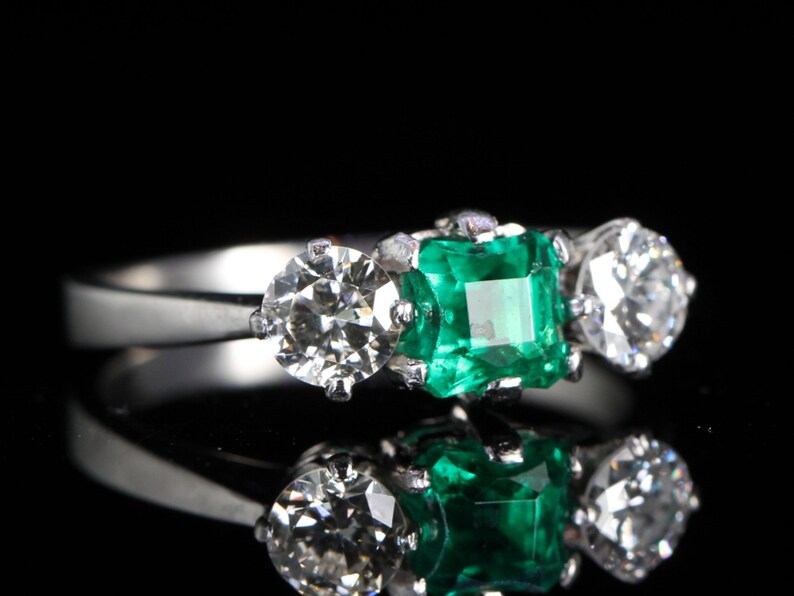 May include: A three-stone ring with a central emerald-cut green gemstone flanked by two round, brilliant-cut diamonds. The ring is set in a silver band, reflecting light against a black background. The ring's design is classic and elegant.