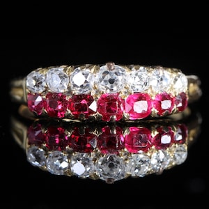 May include: A gold ring with two rows of gemstones. The top row features clear, sparkling stones, while the middle row showcases vibrant red stones. The ring is set against a black background, with its reflection visible.