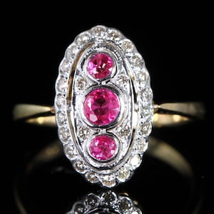 Op de afbeelding: Een ovale ring met een gouden band en een zilverkleurige zetting. De zetting heeft drie ronde, helder roze edelstenen omringd door kleine, heldere stenen. De ring staat tegen een zwarte achtergrond.
