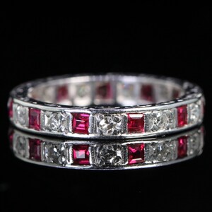 May include: A silver ring with alternating square-cut rubies and clear, round-cut gemstones. The ring is set against a black background, with its reflection visible below. The gemstones are evenly spaced around the band.
