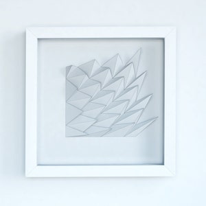 Op de afbeelding: Een wit ingelijst beeld met een wit geometrisch papierontwerp erin. Het ontwerp is gemaakt van veel driehoeken die een 3D-effect creëren.