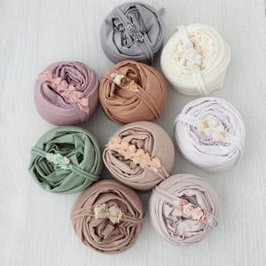 May include: Nine rolled fabric wraps in various pastel colors, each adorned with a matching floral headband. Colors include gray, cream, lavender, peach, sage green, and tan. The wraps are arranged on a light wood-grain surface.