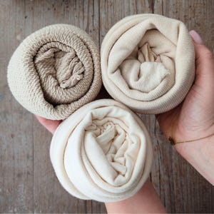 Puede incluir: Tres artículos de tela enrollados en tonos beige y crema se sostienen en una mano. Las telas tienen diferentes texturas, incluyendo un tejido de gofre y un jersey liso. Los artículos están dispuestos en forma triangular sobre un fondo de madera.