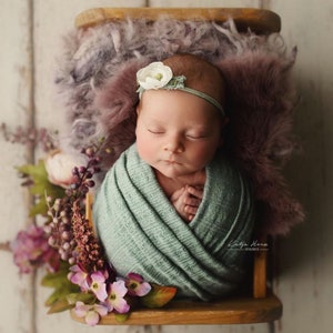 Op de afbeelding: Een pasgeboren babymeisje slaapt rustig in een groene swaddle, gewikkeld in een zacht, pluche deken. Ze draagt ​​een wit bloemenbandje en is omringd door paarse bloemen.