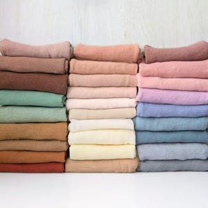 May include: A collection of folded fabric wraps stacked in three vertical columns. The textiles feature a textured, crinkled surface and a diverse color palette including shades of brown, tan, green, pink, blue, and cream.