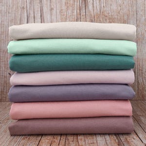May include: A stack of seven folded fabric pieces in various solid colors, including beige, light mint green, dark teal, pale pink, muted purple, dusty rose, and mauve, arranged in a vertical pile with a smooth, matte texture.