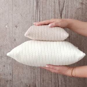 May include: Two knitted pillows, one cream and one off-white, are stacked. The pillows have a textured, ribbed design. The background is a light brown wooden surface.