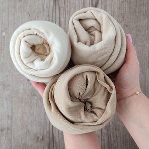 Peut inclure: Trois morceaux de tissu roulés, de couleurs crème, beige et beige clair. Le tissu semble être en maille ou en jersey doux. Les rouleaux sont tenus dans les mains d'une personne, avec une surface en bois en arrière-plan.