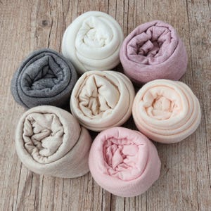 Puede incluir: Siete rollos de tela suave en varios colores, incluyendo blanco, gris jaspeado, beige, melocotón claro y tres tonos de rosa. Cada pieza de tejido está enrollada firmemente en forma circular, mostrando la textura lisa del material.